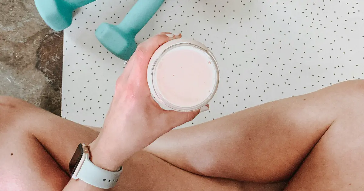 “Person sitting on a workout mat holding a protein shake with colorful dumbbells nearby”